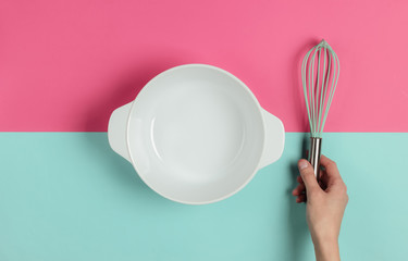 Female hands holding whisk on pink blue background with empty ceramic bowl. Top view