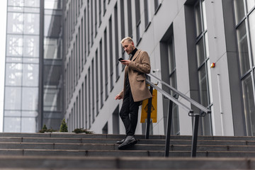 Fototapeta premium Handsome businessman standing in a beige coat with crossed legs and looking at the phone.