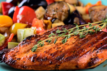 Appetizing grilled chicken fillet with a sprig of thyme is on a ceramic plate next to various fryed vegetables, macro