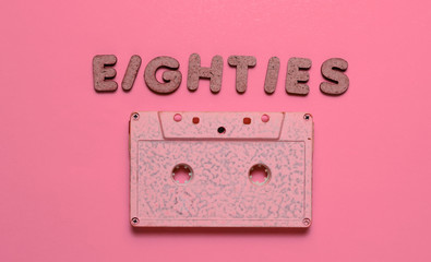 Creative retro style concept, 80s. Audio cassette on pink background with the word Eighties from wooden letters, minimalism
