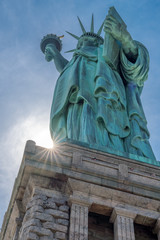 Obraz premium Statue of Liberty with sunburst. Image of the Statue of Liberty shot from a unique angle: looking up and timed so that a partially obscured sun makes a starburst at the base of the pedestal.