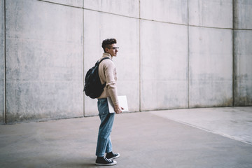 Young man in glasses carrying laptop near grey wall