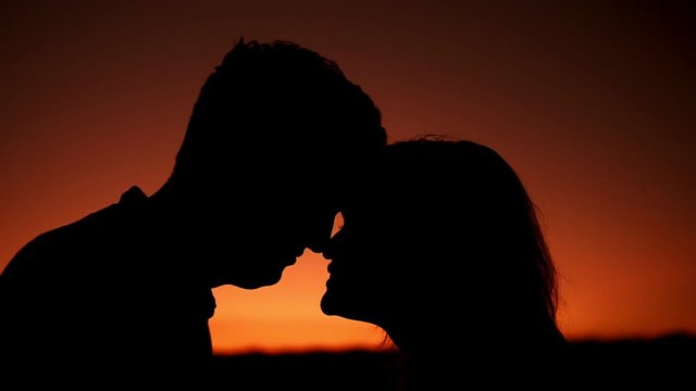 Silhouettes of lovers head to head on sunset background. Theme of love and happiness of family. A man and a woman they look at each other against background of orange gradient of sunset.