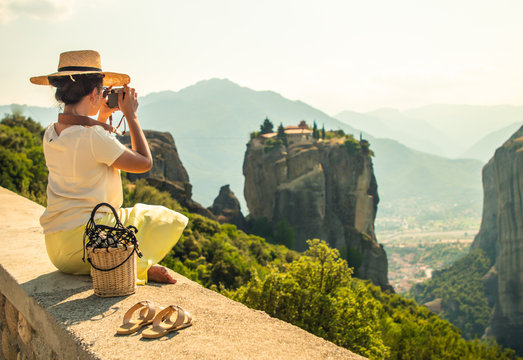 Young woman with white dress and large hat sitting in front of greece meteor mountains, monastery and village in the background