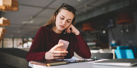 Pondering young female blogger in cafe creating new publication for social networks via smartphone, beautiful hipster girl holding mobile phone in hands reading latest news from browsed websites