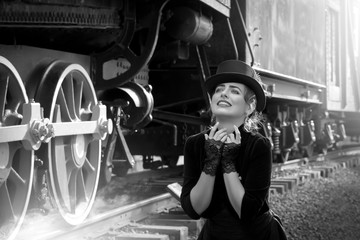 Beautiful girl in a black dress and hat near an old steam locomotive and big iron wheels. Blond beauty. Vintage portrait of the last century, retro journey.