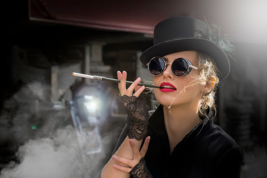Steampunk girl in a black dress and hat near an old steam locomotive and large iron wheels. Blond beauty. Vintage portrait of the last century, retro journey.