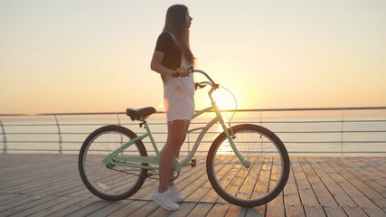 Fototapeta na wymiar Young beautiful woman with a bicycle is having a good time at the sea at sunset or sunrise 