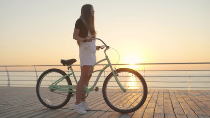 Young beautiful woman with a bicycle is having a good time at the sea at sunset or sunrise 