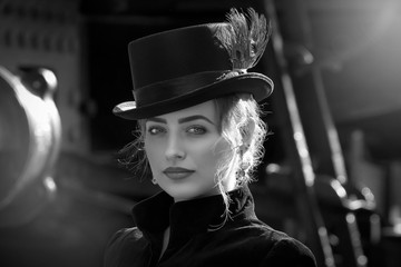 Beautiful girl in a black dress and hat near an old steam locomotive and big iron wheels. Blond beauty. Vintage portrait of the last century, retro journey.