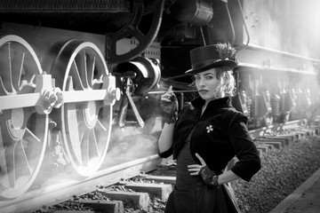 Beautiful girl in a black dress and hat near an old steam locomotive and big iron wheels. Blond beauty. Vintage portrait of the last century, retro journey.