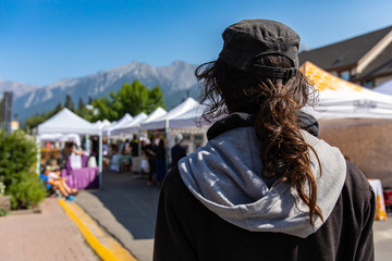 Fototapeta A man wearing a dark hooded top and baseball cap is seen from behind, looks towards a busy street filled with market stalls, with copy space on left obraz