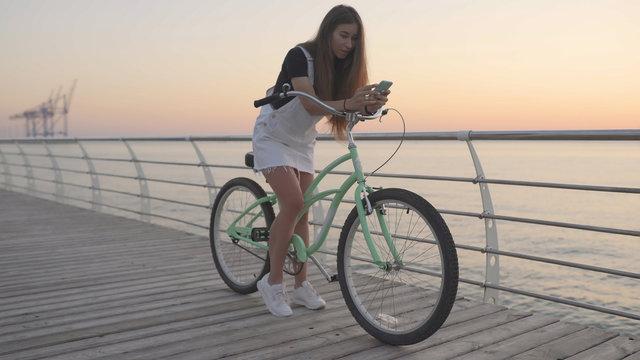 Young Attractive Woman Uses A Smartphone And Riding Vintage Bike Near The Sea During Sunrise Or Sunset