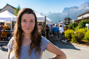 A close up and front portrait of a confident caucasian girl in early thirties with pierced nose, standing by a blurry street scene during local fair