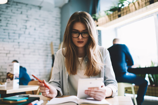 Attractive young woman received message on gadget checking notification, beautiful skilled hipster girl in eyeglasses reading news via modern device at cafeteria and writing article for share