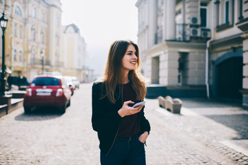 Young positive trendy smiling blogger looking aside while waiting friend outdoors and creating new publication for social network, happy hipster girl holding mobile phone in hand and checking mail