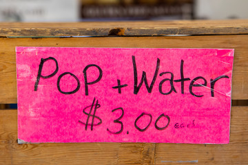 Obraz premium A colorful pink sign is seen close up, fixed to a wooden crate, advertising pop and water for sale on a market stall at a local farmers fair