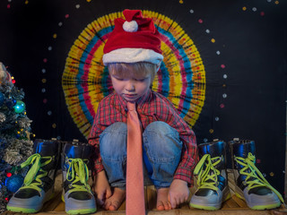 charming little boy is sitting in festive attire next to luminous and blinking Christmas lights. Glasses reflect flashing garlands with lights. Christmas tree, colored background, santa claus, child