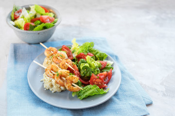 tiger prawn skewers on cauliflower risotto with salad of lettuce and tomatoes, plate and bowl on a blue napkin, light gray stone surface with copy space, selected focus, narrow depth of field