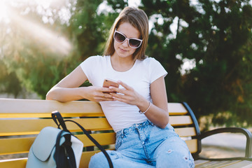 Obraz premium Attractive teenage hipster girl in sunglasses sitting on bench and communicated with friends via smartphone application, beautiful woman searching touristic tour connected to 4g internet in roaming