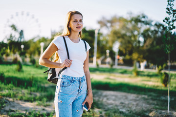 Portrait of successful smiling young hipster girl 20s walking and enjoying summer weather, positive blonde woman strolling while looking at camera and holding modern smartphone at sunny spring day