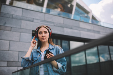 Attractive trendy hipster girl in jeans jacket enjoying listening music via modern earphones while holding new mobile phone in hand and looking away, thoughtful young female meloman outdoors