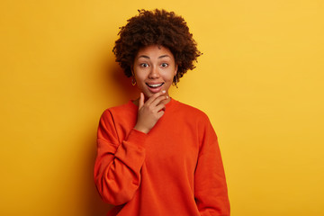 Fototapeta na wymiar Cheerful curly haired woman holds chin, expresses bright emotions, reacts on unexpected news, learned something surprising, likes what she hears, wears casual jumper, poses over yellow wall.