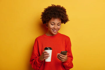 Smiling beautiful dark skinned woman holds smartphone, plays interesting game online, sends meme, drinks takeaway coffee, wears casual jumper, has Afro hairstyle, isolated on yellow background