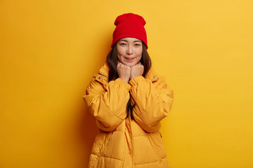Portrait of beautiful gorgeous female feels shy, keeps hands under chin, wears red hat and winter coat, looks directly at camera with gentle smile, isolated over yellow background. Seasonal fashion