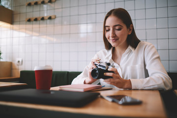 Caucasian positive female photographer 20s holding retro camera in hands and checking images sitting in coffee shop, young amateur hipster girl happy from spending time for favourite hobby in cafe