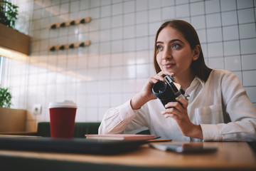 Attractive dreaming hipster girl 20s think about profession photograph while holding vintage camera in hands and looking away, young smiling woman with retro technology resting at coffee shop