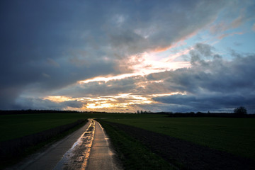 Wet reflecting country road made of concrete slabs is leading through the dark fields under a dramatic cloudy sky with evening sun, rural landscape in Mecklenburg, Germany, copy space