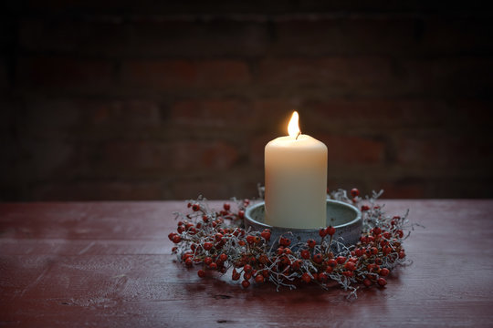 Burning candle in a bowl with a small wreath from rose hips and silver cushion bush on a red wooden table, winter decoration for Advent, Christmas and New Year, dark background with copy space