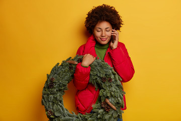 Portrait of happy pleased curly African American woman carries Christmas wreath, enjoys speaking...