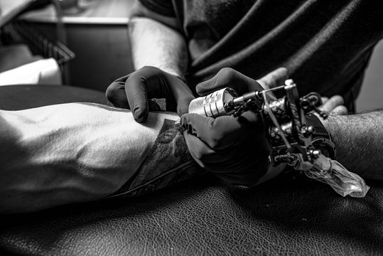 Male arm with tattoo. Master in black gloves is preparing to draw new tattoo. Tattoo machine. Black and white.
