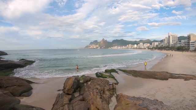 Sideways pan showing early morning sunrise with waves coming in at an almost empty Ipanema beach against a blue sky with clouds and Two Brothers mountain of Leblon in the background