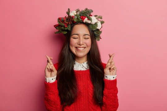 Making Christmas wish concept. Happy brunette girl stands with fingers crossed, bites lips and awaits miracle happen, wears green decorative spruce wreath, red knitted jumper, believes in good luck