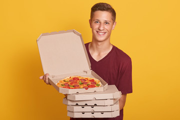 Photo of happy joyful man wearing burgundy casual t shirt, looking smiling at camera and holding pizza boxes, keppeng top box opened, posing isolated over yellow background. Delivery concept.