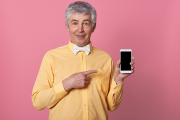 Indoor shot of mature grey haired man in yellow shirt with bow tie, holding mobile phone in hands and pointing at black blank screen, looks directly at camera, isolated over pink studio background.