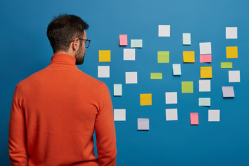Back view of male freelancer in orange turtleneck concentrated on sticky notes with blank space...