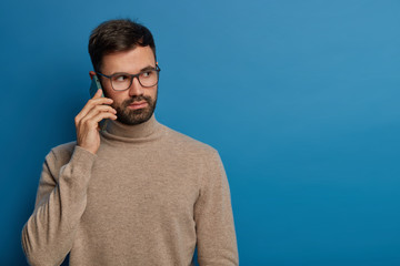 Photo of serious bearded young guy talks on phone, calls someone via modern gadget, expresses...