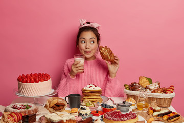 Addicted young Asian female drinks milk and eats delicious croissant, licks lips from pleasant taste, enjoys desirable favourite dessert, sits at table with many sweet products, isolated on pink wall