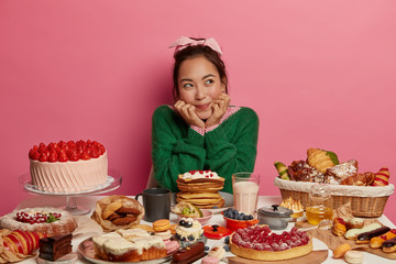 Unhealthy nutrition and much calories concept. Thoughtful brunette Asian woman poses at festive table, looks aside, has doubts eat or not, surrounded with many bakery products, wears green jumper