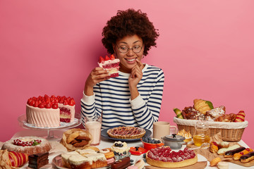 Photo of happy curly haired female holds big piece of strawberry cake, eats yummy desserts on her...