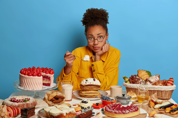 Horizontal shot of sad dark skinned woman eats cream from pancakes, eats holiday treats, feels...