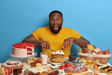 Amazed dark skinned man eats pancakes, uses fork and knife, wears yellow t shirt, has delicious...