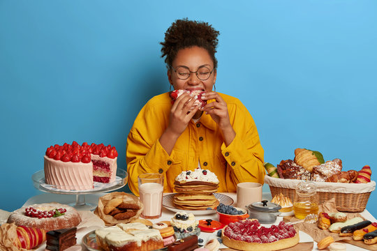 Cheat meal and gluttony concept. Ethnic curly woman eats strawberry creamy cake with much calories, has sweet lunch, tastes various desserts, leads sweet life with confectionery. Female fan of bakery