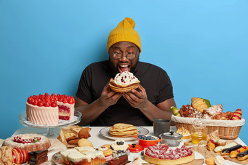 Happy celebrating sweet tooth man bites baked pancakes with cream, eats all desserts by himself, doesnt prevent from obesity, makes wrong nutrition choice, greedily devours bakery production