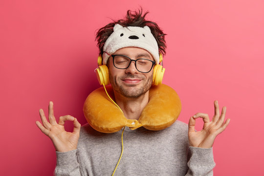 Pleased guy makes oaky gesture with both hands, meditates during listening music, has unforgettable journey, uses neck pillow for travelling on long distance, feels relaxed. People, journey, yoga, nap