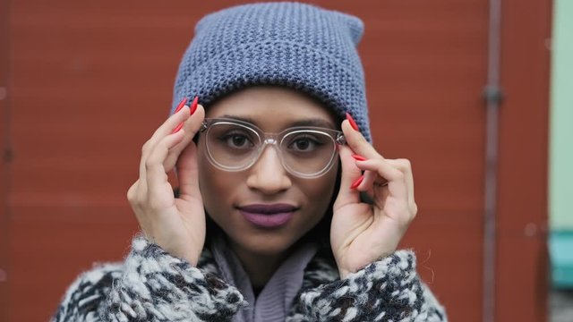 Millennial woman in glasses looking at camera and smiling. Female in hat and coat outdoors. Close-up portrait of beautiful mixed race woman in 4K, UHD